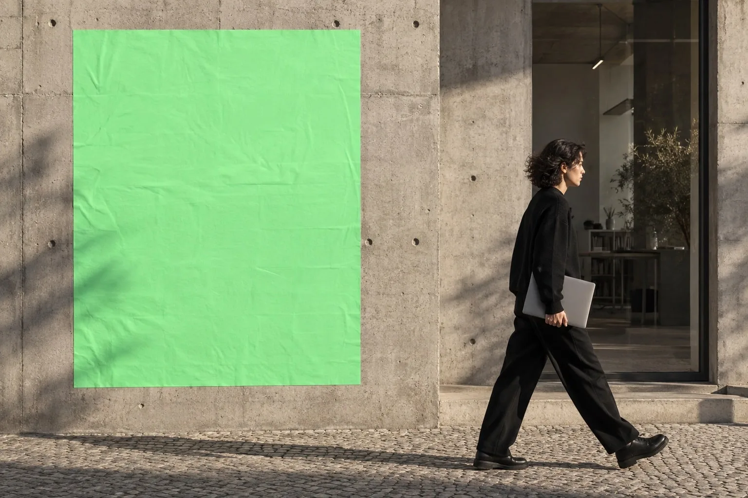 A person with a laptop walks past a green poster on a concrete wall.